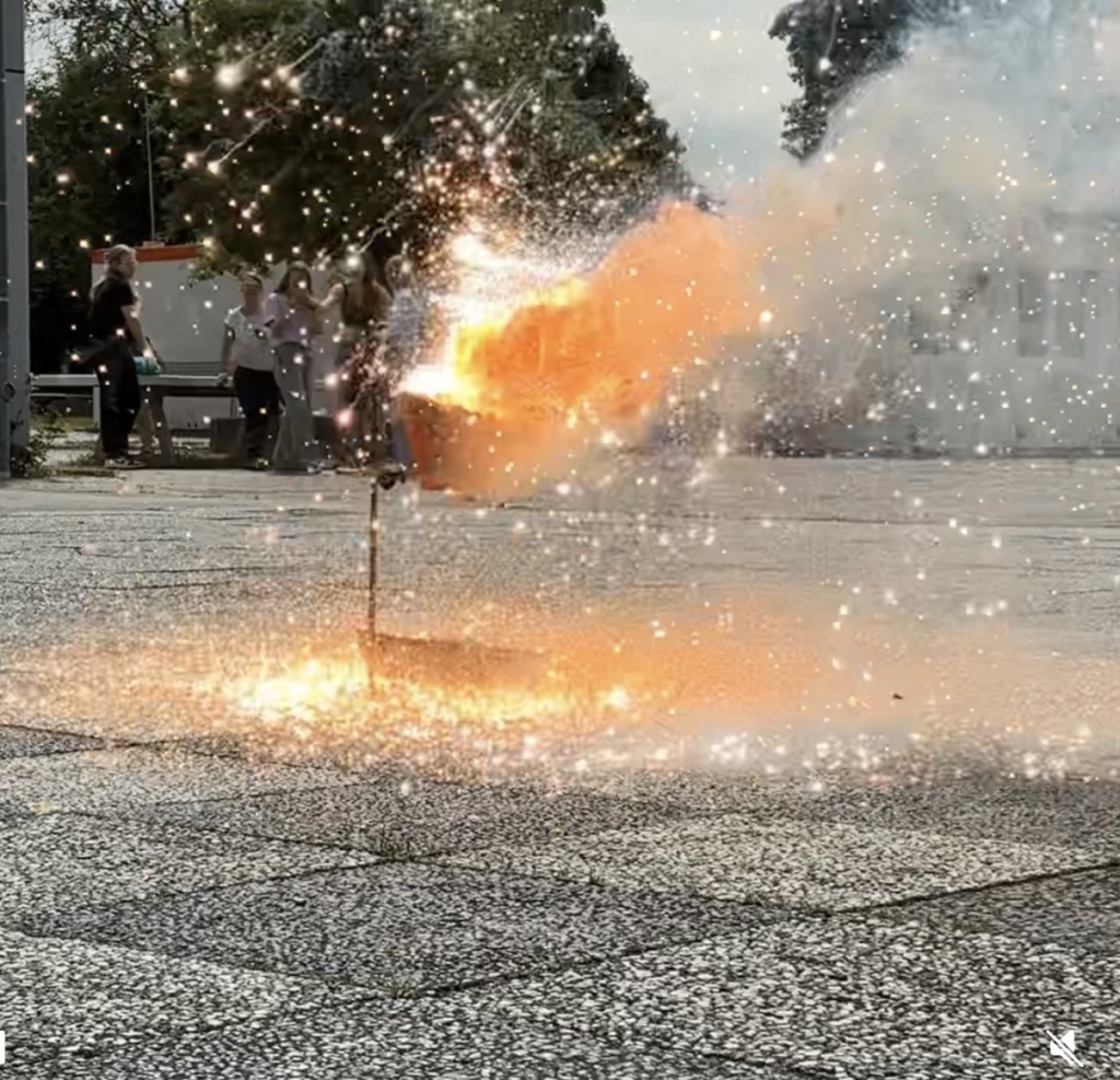 Screenshot Auf dem Schulhof entwickeln sich an einem Metallstab Rauch und Funken, ein chemisches Experiment.