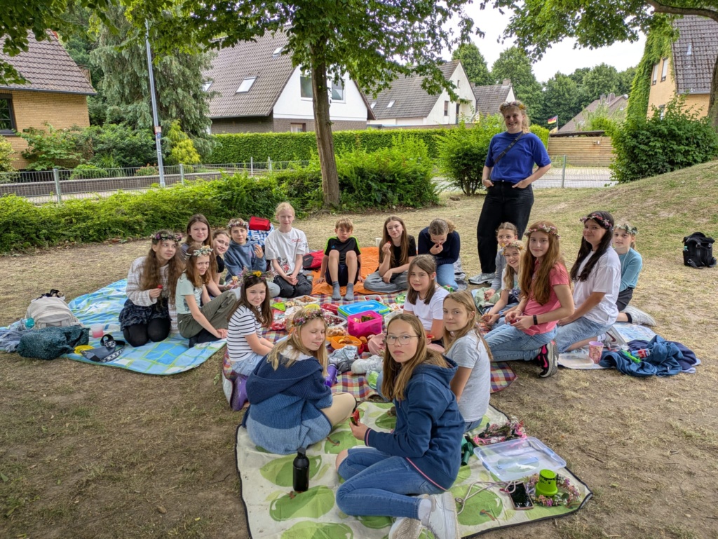 Midsommer1 Circa 30 Schülerinnen und eine Lehrerin picknicken im Grünen mit Blumenkränzen im Haar.