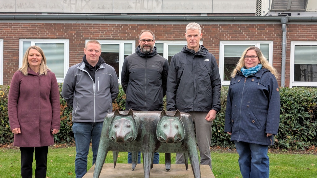 Drei Männer und zwei Frauen stehen freundlich um eine grünen Doppelwolfplastik herum, im Hintergrund Teile des GGI.