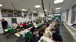 IMG-20251202-WA0013 Ca. 50 Schüler sitzen sich an drei langen Tischreihen gegenüber mit einem Schachbrett in der Mitte, eine Lehrkraft steht am Rande.