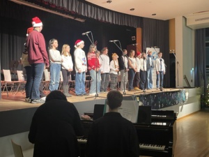 IMG_7393 Ein gemischter Schülerchor mit Weihnachtsmützen singt auf einer Bühne begleitet von Klavierspielern.