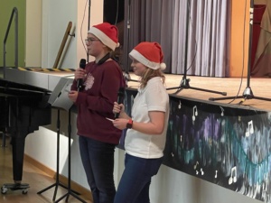 IMG_7403 Zwei Schülerinnen mit Weihnachtsmützen machen vor der Bühne eine Ansage.