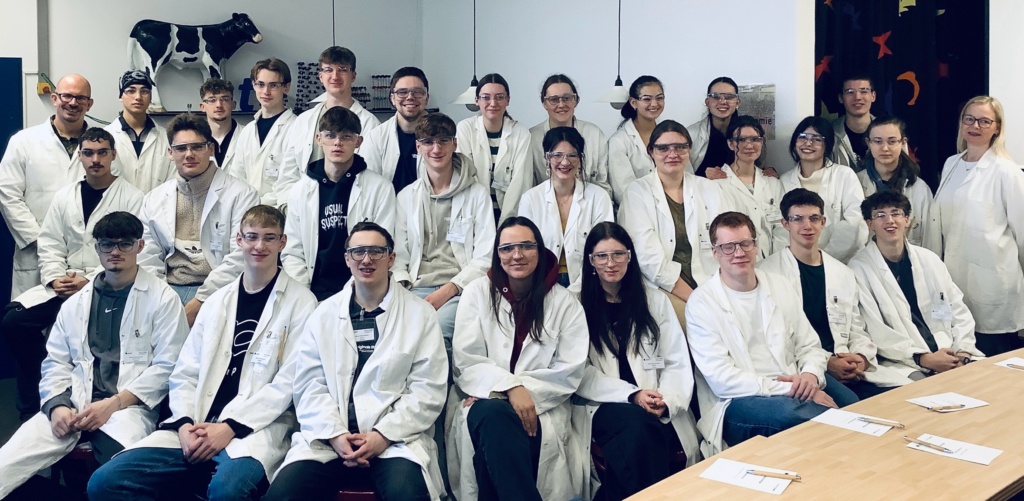 Cat. 30 junge Erwachsene sitzen in drei Reihen hintereinander und tragen weiße Laborkittel, sie lächeln in die Kamera.