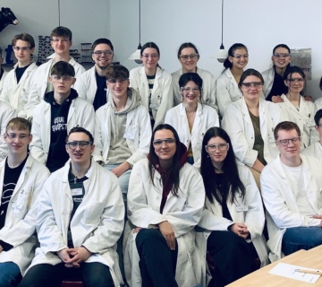 Cat  30 junge Erwachsene sitzen in drei Reihen hintereinander und tragen weiße Laborkittel, sie lächeln in die Kamera 