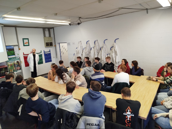 An Tischen n U-Form sitzen ca. 30 Schülerinnen und Schüler und hören einem Mann in weißem Laborkittel zu